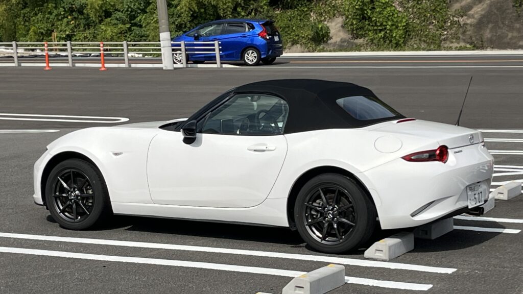 幌を閉じた白のNDロードスターを横から見たところ。奥に青いコンパクトカーが停まっている